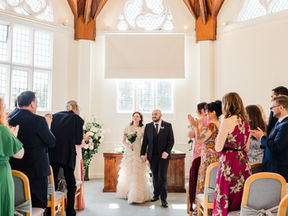 Bride and groom walk hand in hand down the aisle in sunlit Octagonal Suite at Cheshunt Registry Office as guests cheer and applaud. Floral details and wooden beams visible.