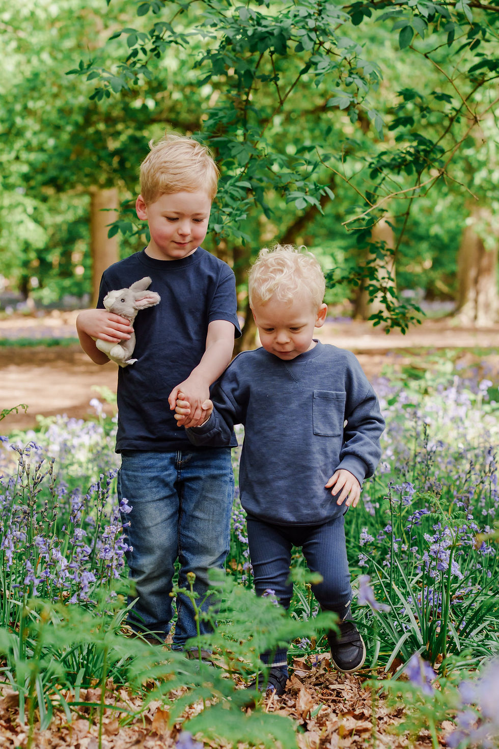 Two young boys in a forest clearing. One holds a stuffed rabbit, guiding the other by hand. Surroundings are lush with green leaves.