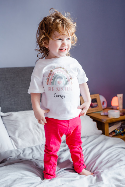 Smiling girl standing on a bed wearing a t-shirt with a rainbow motif and 'Big Sister' lettering