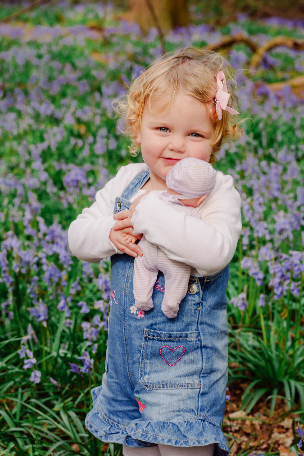 Young girl with blonde curls and pink bow, holding a toy doll. She's wearing denim overalls, standing in a field of purple flowers.