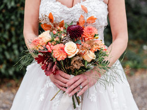 Bride in a lace dress holds a colourful bouquet of orange and cream flowers with berries and autumn leaves. Greenery background. Warm, romantic mood.