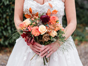 Bride in a lace dress holds a colourful bouquet of orange and cream flowers with berries and autumn leaves. Greenery background. Warm, romantic mood.