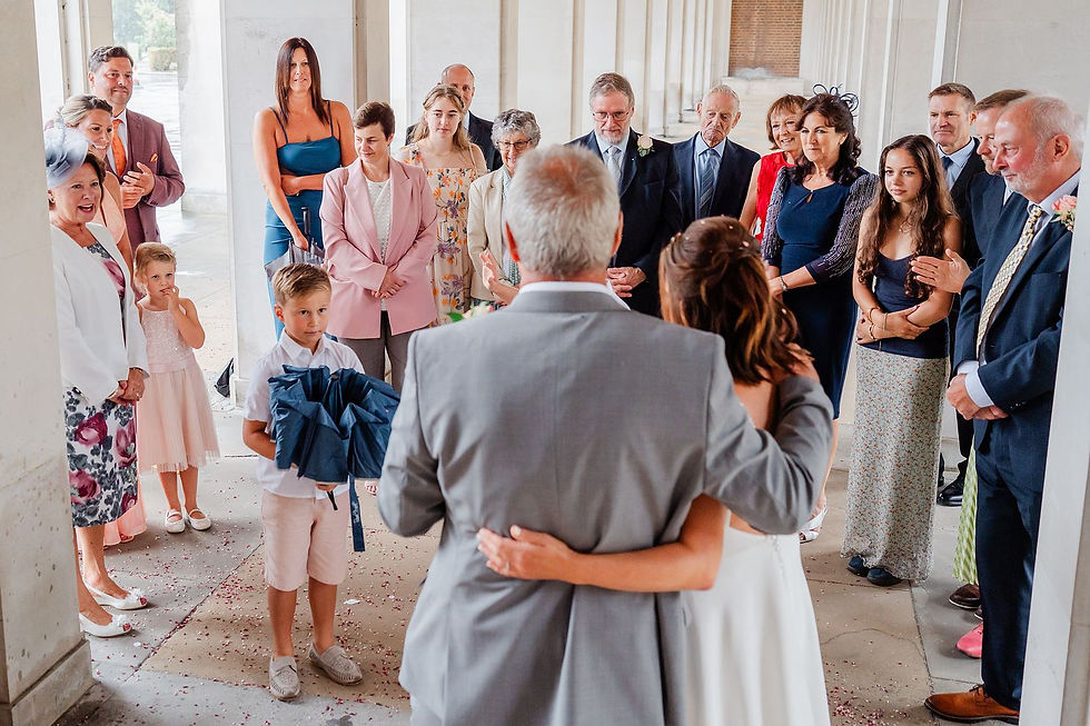 A wedding couple embraces in a hall surrounded by guests wearing colourful attire. A boy holds umbrellas. The mood is joyful and celebratory.