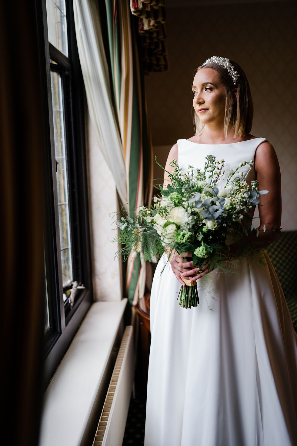 Hanbury Manor wedding. Bride holding green and cream themed bouquet and gazing out of the window