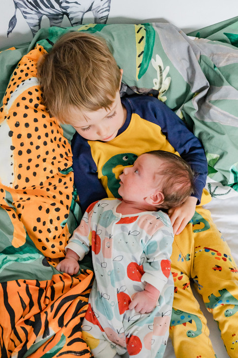Young boy in yellow pyjamas sitting on his bed with a newborn baby propped up on his lap