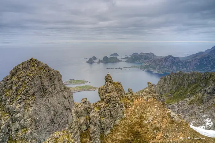 Dramatiske fjelltopper i Norge med hav og øyer under grå skyer.