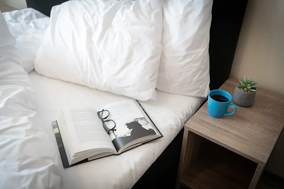 Åpen bok, briller på ren hvit seng med kaffe og plante.