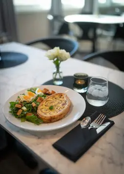 Quiche med salat, vann, bestikk og blomster på bordet.