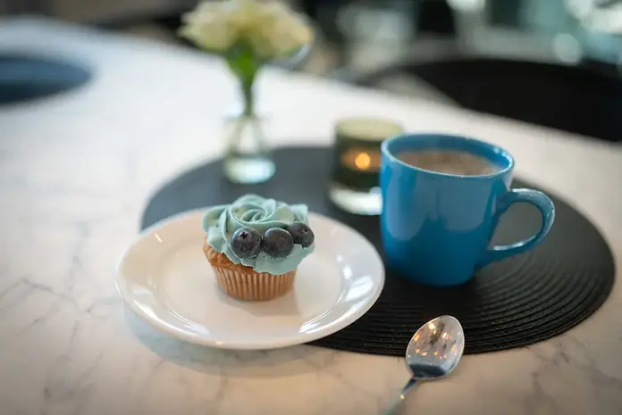 Velsmakende blå cupcake, kaffekopp, tent lys, blomster på hvit marmor.