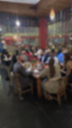 A lively group of people sit around a wooden table in a warmly lit restaurant, engaged in conversation. The space is decorated with hanging ornaments, creating a festive atmosphere.