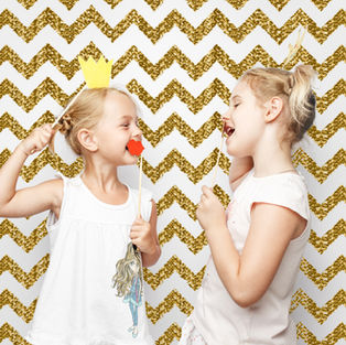 Two girls laughing, holding stick props: a crown and red lips. Gold chevron background. Bright, playful mood.