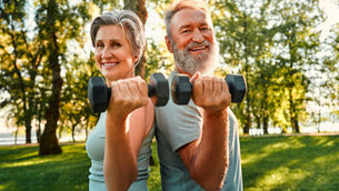 Ouder stel traint samen met dumbbells in een park, lachend en fit terwijl ze rug aan rug staan in de buitenlucht.