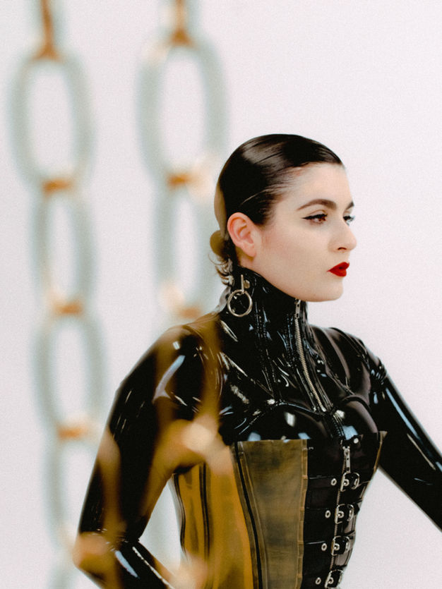 3/4 shot of Boston & NYC Professional Dominatrix Mistrix Violeta Félix wearing a shiny latex catsuit layered with a natural translucent colored and black latex Demask corset with silver buckles on the front and a black pvc neck corset with silver rings on the side.  