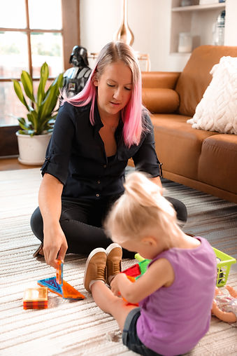 A female therapist with blonde and pink hair playing with a child who is sitting on the floor with several toys surrounding her in a therapy room.