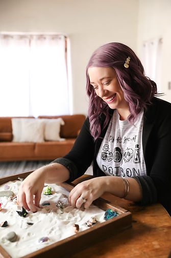 A female therapist with purple hair, grey t-shirt and black cardigan smiling with her hands in a sand tray with several miniature figures inside.