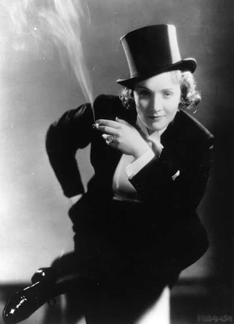 Black-and-white portrait of a woman in a top hat and tuxedo, smoking a cigarette with a sly, confident pose against a plain backdrop.