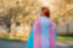 Trans right movement. A person with red hair draped in a transgender pride flag stands outdoors. Autumn trees and a building are in the background.