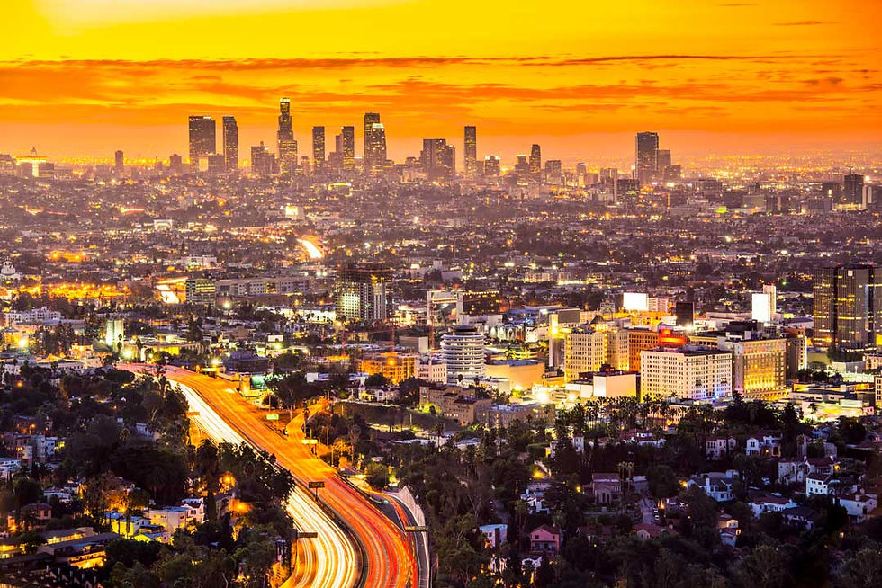 Orange sunset over Los Angeles skyline, with glowing highway light trails and city lights stretching into the distance.