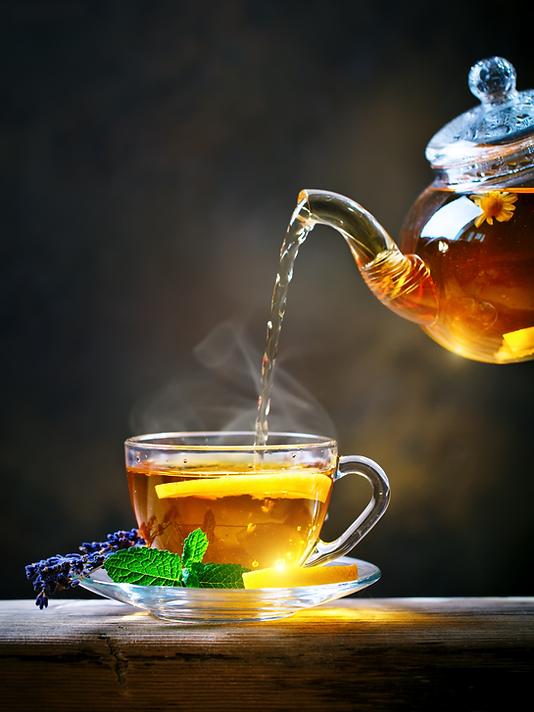 Glass teapot pouring amber tea into a steaming cup with lemon, mint, and lavender on a saucer against a dark background