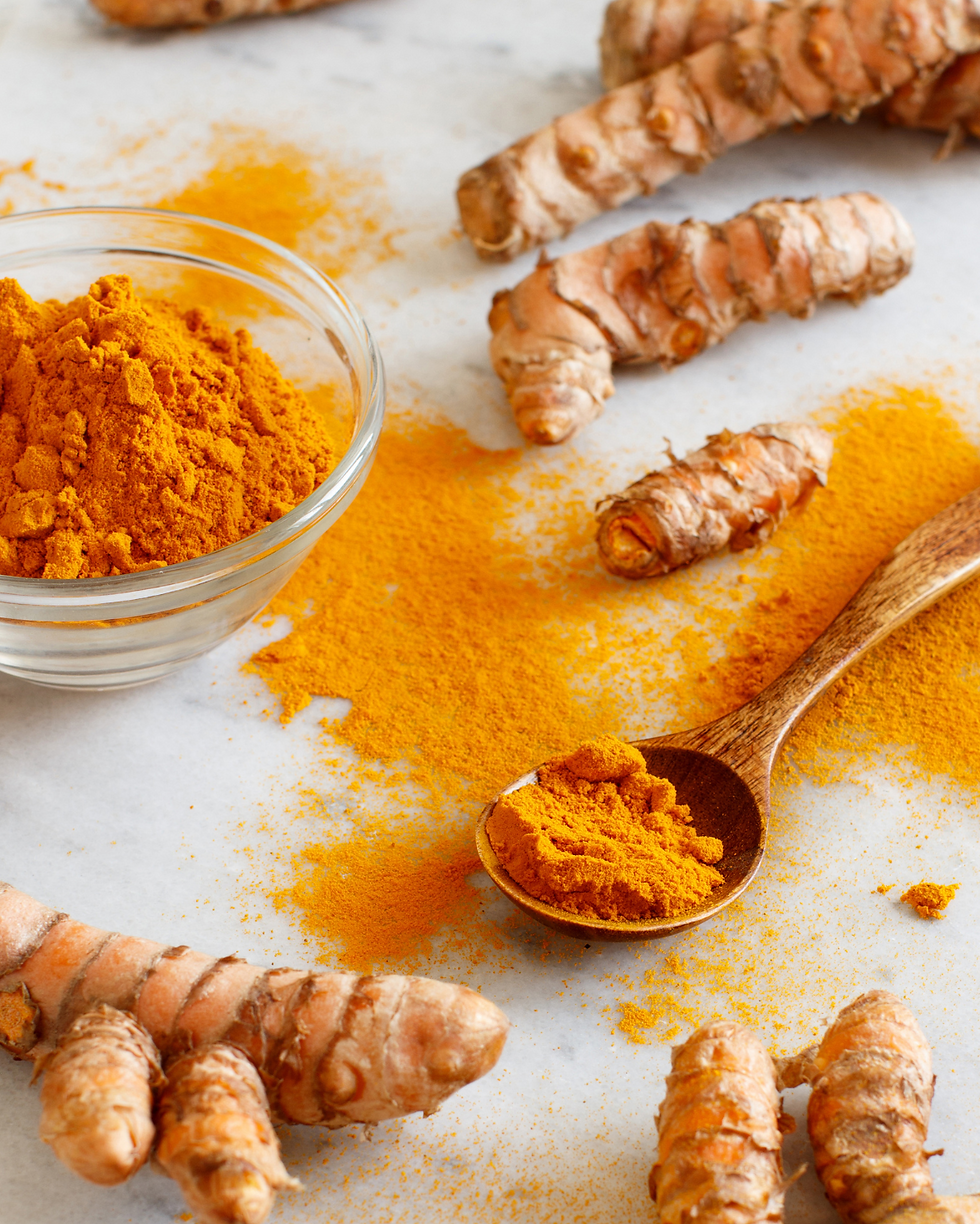 Turmeric roots surround a bowl and wooden spoon of vibrant orange turmeric powder on a light marble surface, creating a rich, earthy feel.
