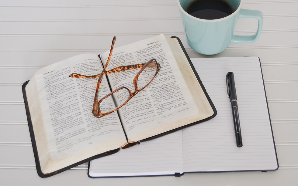 An open Bible with glasses resting on its pages, accompanied by a notebook, pen, and a mug of coffee on a white wooden table,