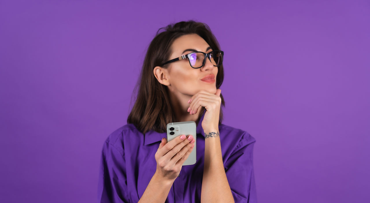 young-woman-shirt-glasses-purple-background-with-phone-looks-thoughtfully-side-with-slight