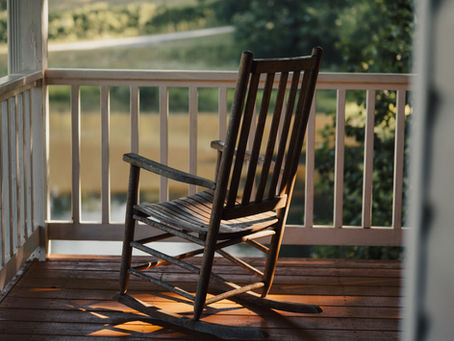 An empty rocking chair on a quiet front porch overlooking fields at sunset, inviting reflection after a day's work.