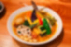 Colorful vegetable curry in a white bowl on a wooden table, topped with lotus root, baby corn, peppers, squash, and beans.