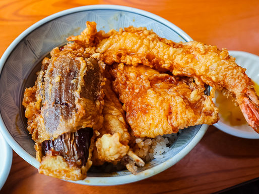 Bowl of tempura shrimp and eggplant over rice on a wooden table, with a small dipping dish beside it.