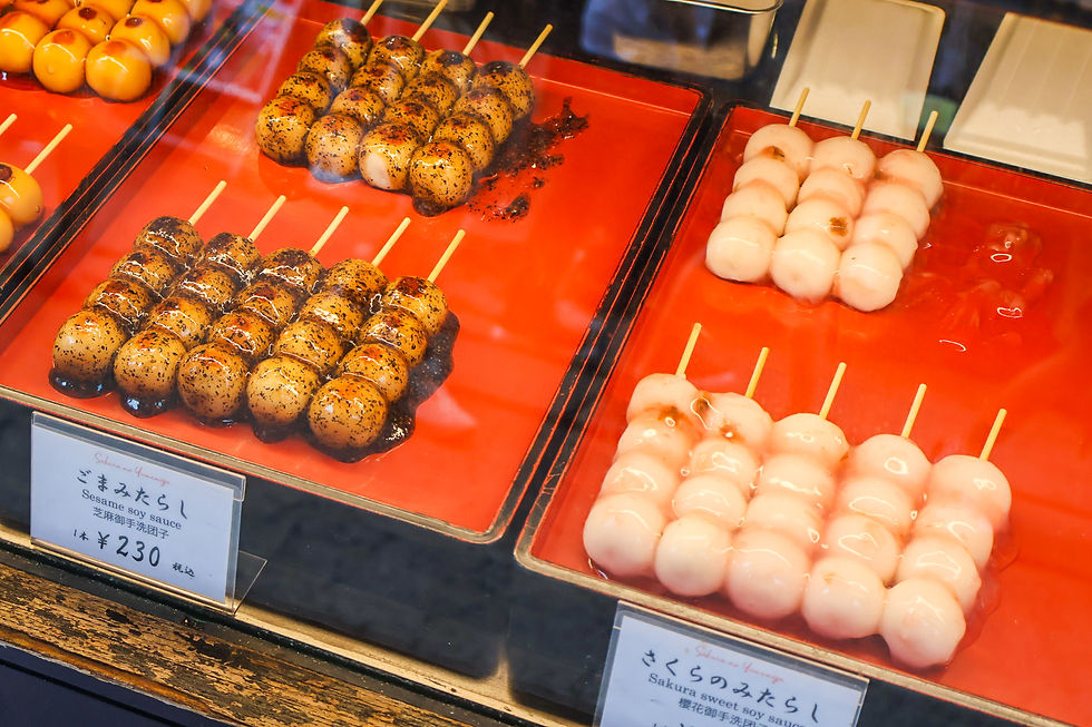 Skewers of glossy dango in a red display case, some coated in sesame soy sauce and others pale pink, with price labels.