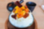 Shaved ice dessert topped with glazed pineapple chunks, pecans, and syrup on a wooden tray, with black cups in the background.
