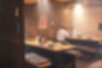 Two men talk over a meal in a warm, dim Japanese restaurant with wooden tables, grill inserts, and a poster on the wall.