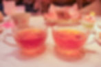 Two ornate glass cups with pink tea on a marble table. Background includes floral decor, pastries, and layered desserts. Cozy ambiance.
