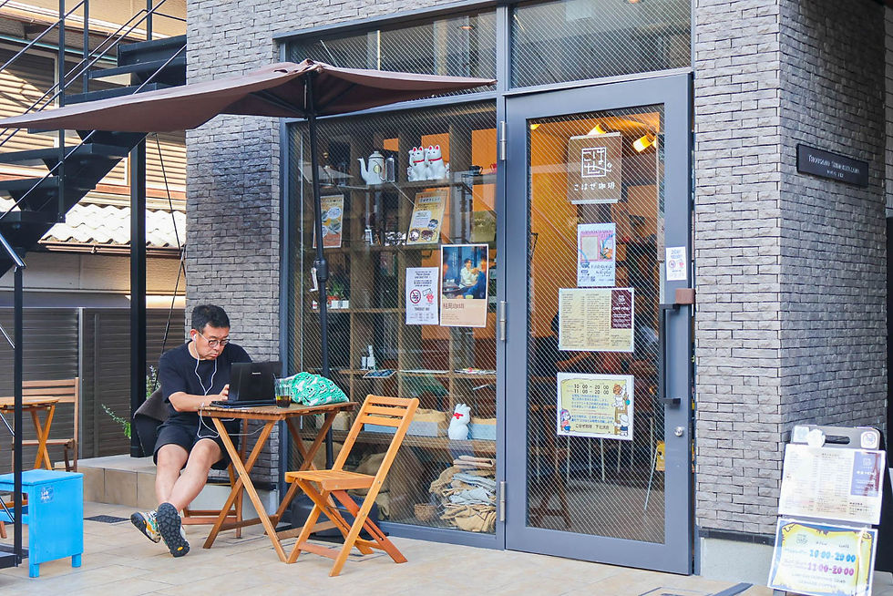 Man with earbuds works on a laptop at an outdoor café table beside a glass storefront with Japanese signs and a large umbrella.