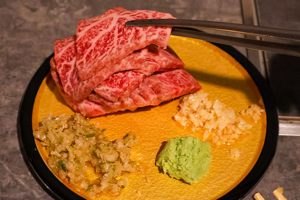 Chopsticks lift marbled raw beef over a yellow plate with rice, wasabi, and chopped onions on a table.