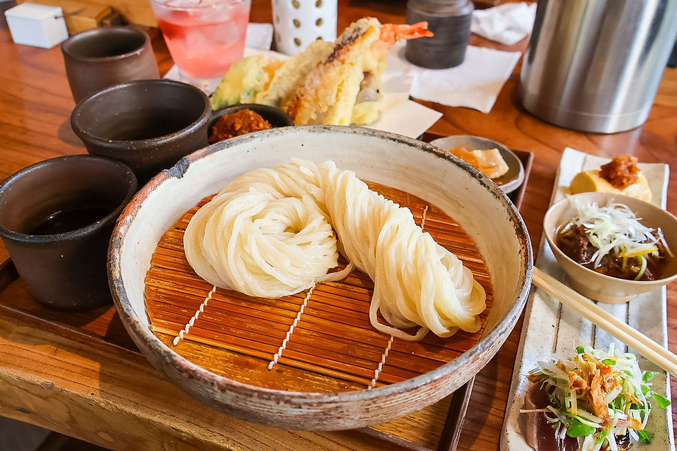 Japanese noodle dish in a ceramic bowl with tempura, dipping sauces, and side dishes on a wooden table.
