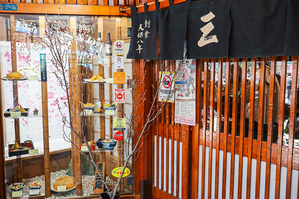 Japanese restaurant entrance with food sample display, black noren curtains, cherry blossom branches, and Japanese signs in warm wood setting