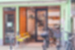 Japanese ramen shop storefront with glass door, yellow stools, parked bicycles, and Japanese signs in a mint-green building.
