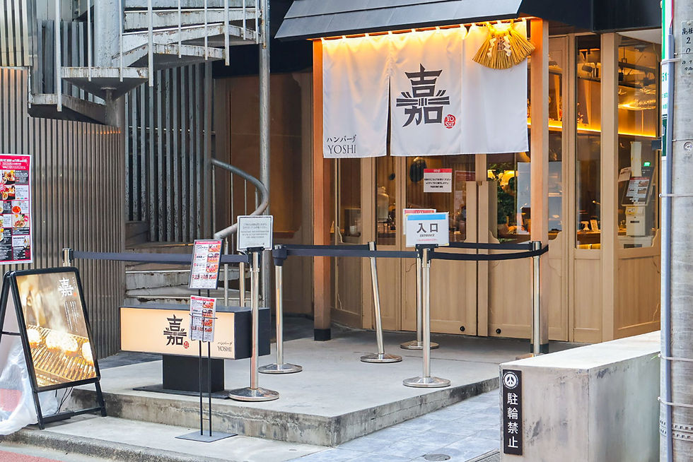 Warm-lit Japanese restaurant entrance with white noren reading YOSHI, menu signs, and queue stanchions on a quiet street.