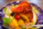 Slice of fruit-topped cake on a blue plate with kiwi, grapes, mint, and a fork in a warm, restaurant-like setting.