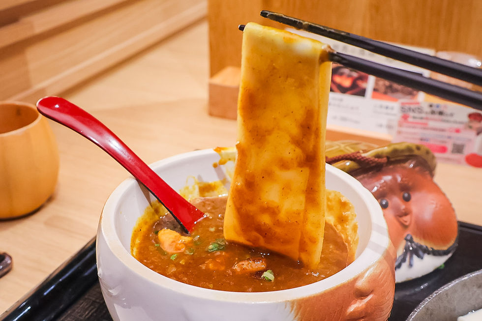 Chopsticks lift a wide noodle sheet from a bowl of spicy ramen beside a red spoon on a wooden table.