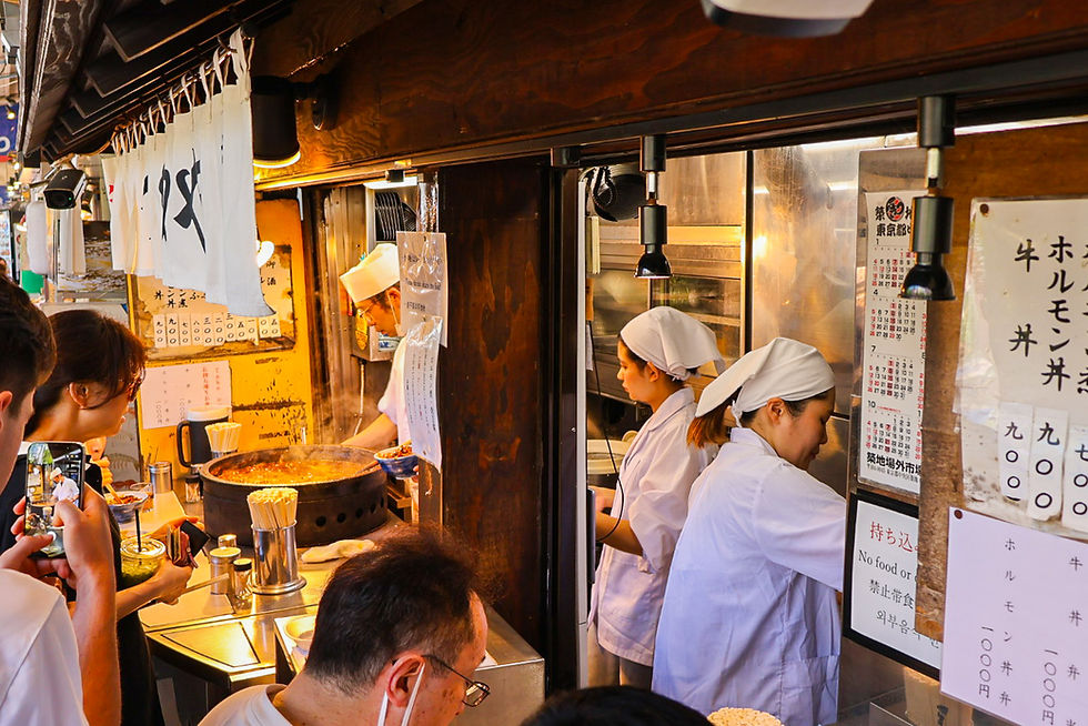 Busy Japanese food stall with chefs in white hats serving steaming dishes to customers; handwritten menus and signs on the counter.