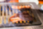 Two pieces of meat sizzling on a smoky metal grill, with blue flames and charred edges.