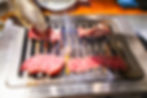 Raw beef slices grilling on a metal tabletop grill, tongs lifting one piece as flames and smoke rise in a warm restaurant setting