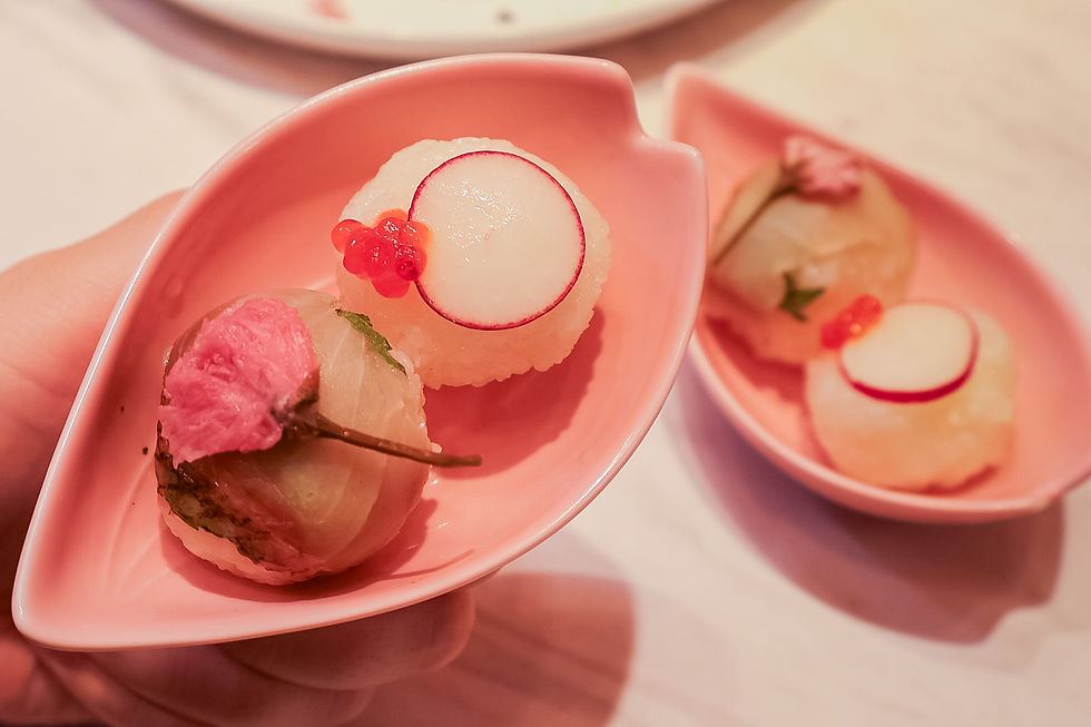 Sushi balls garnished with radish and salmon roe on pink leaf-shaped plates. A hand holds one plate. Subtle, elegant dining ambiance.