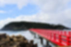 Bright red bridge stretches over waves to a forested island, with concrete wave breakers in the foreground and cloudy blue sky.