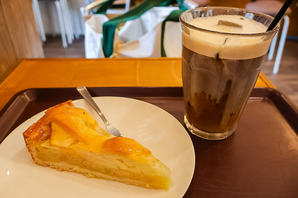 Slice of apple pie on a white plate with a spoon, beside a glass of iced coffee on a tray in a cozy café.