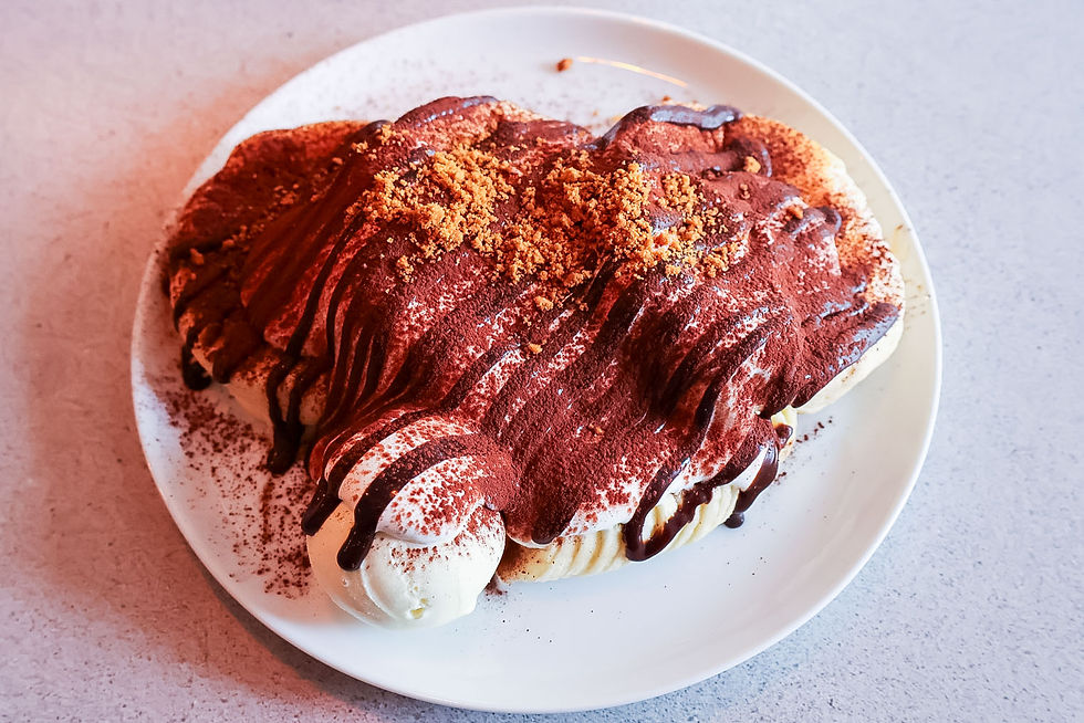 Tiramisu-like dessert on a white plate, dusted with cocoa and cookie crumbs, drizzled with chocolate sauce on a pale tabletop.