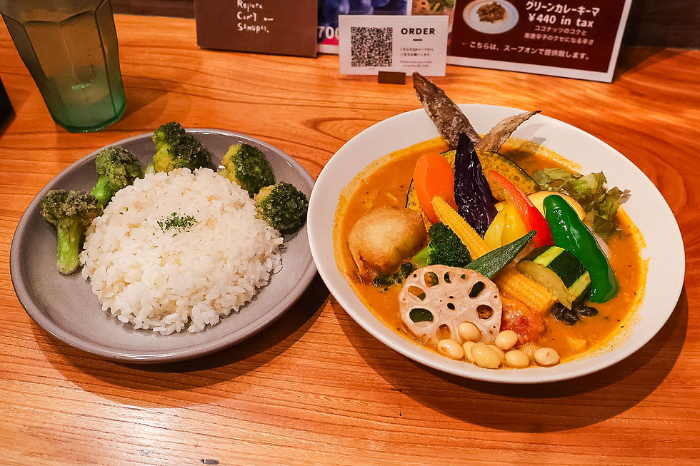 Japanese curry with colorful vegetables and beans in a white bowl, plus rice and broccoli on a gray plate, on a wooden table.
