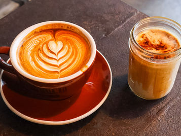 Latte art coffee in a red cup and a cinnamon-topped iced coffee in a jar on a dark tabletop.